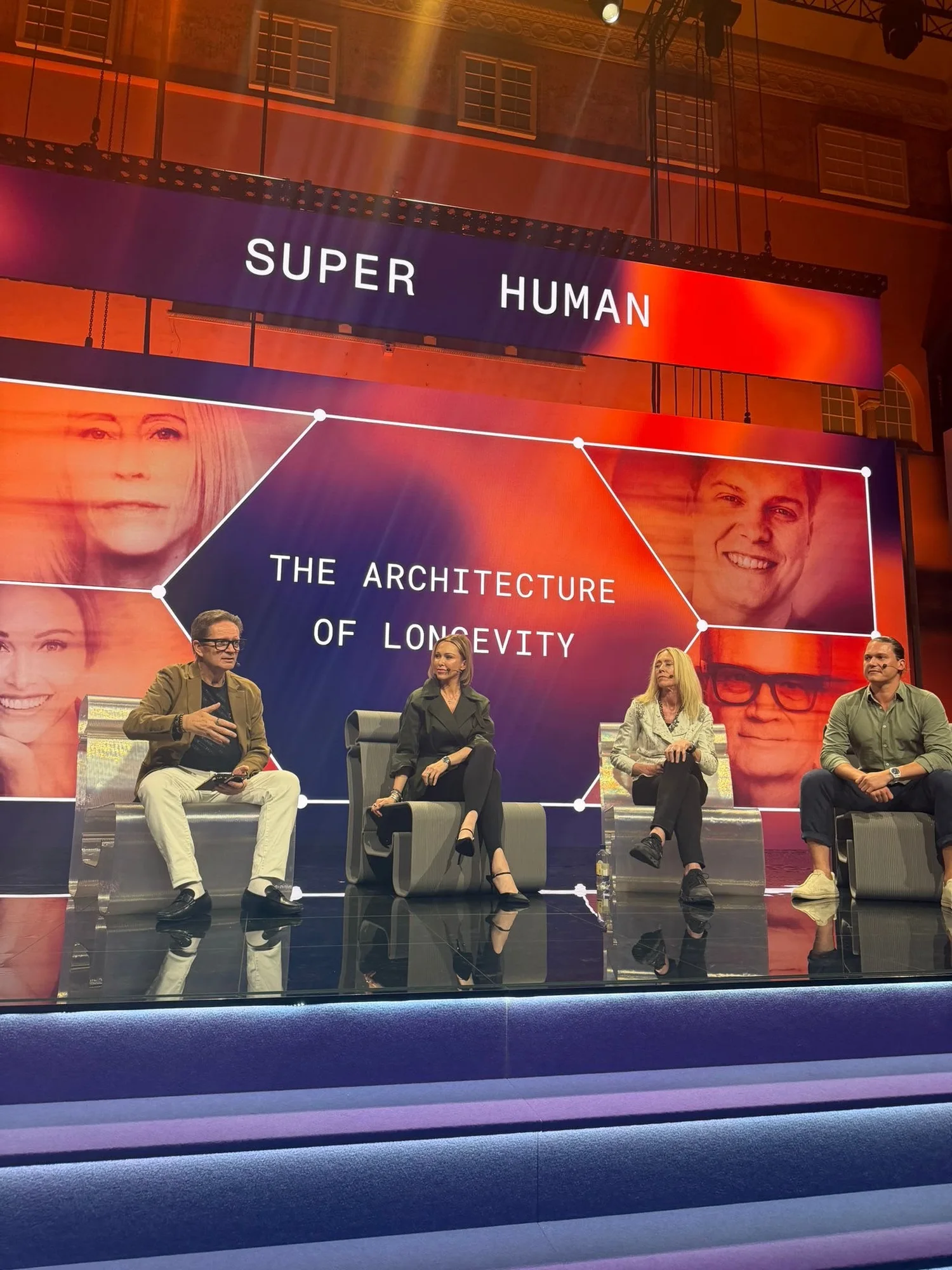 The Architecture of Longevity panel, four speakers seated across the stage beneath the title on the screen behind them