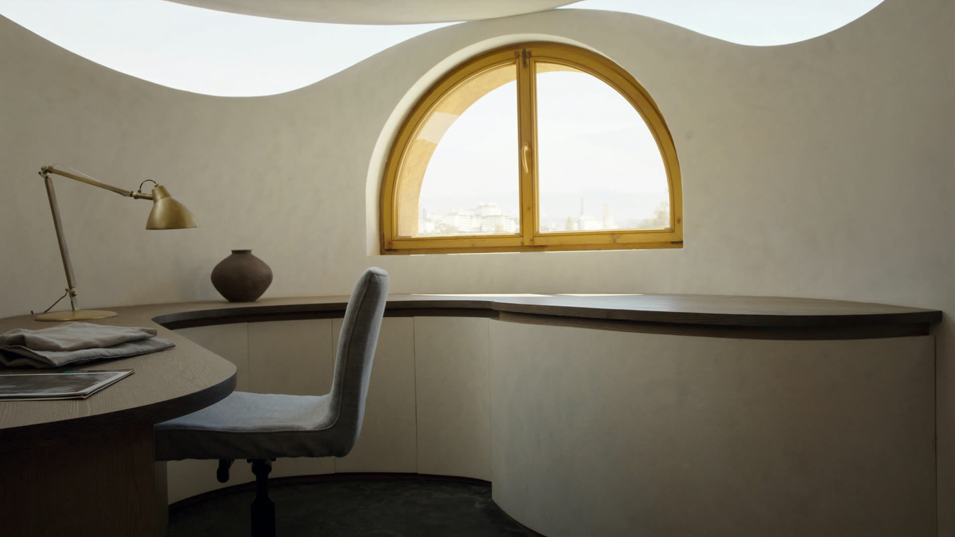 A curved oak desk under an arched clerestory window, brushed brass lamp