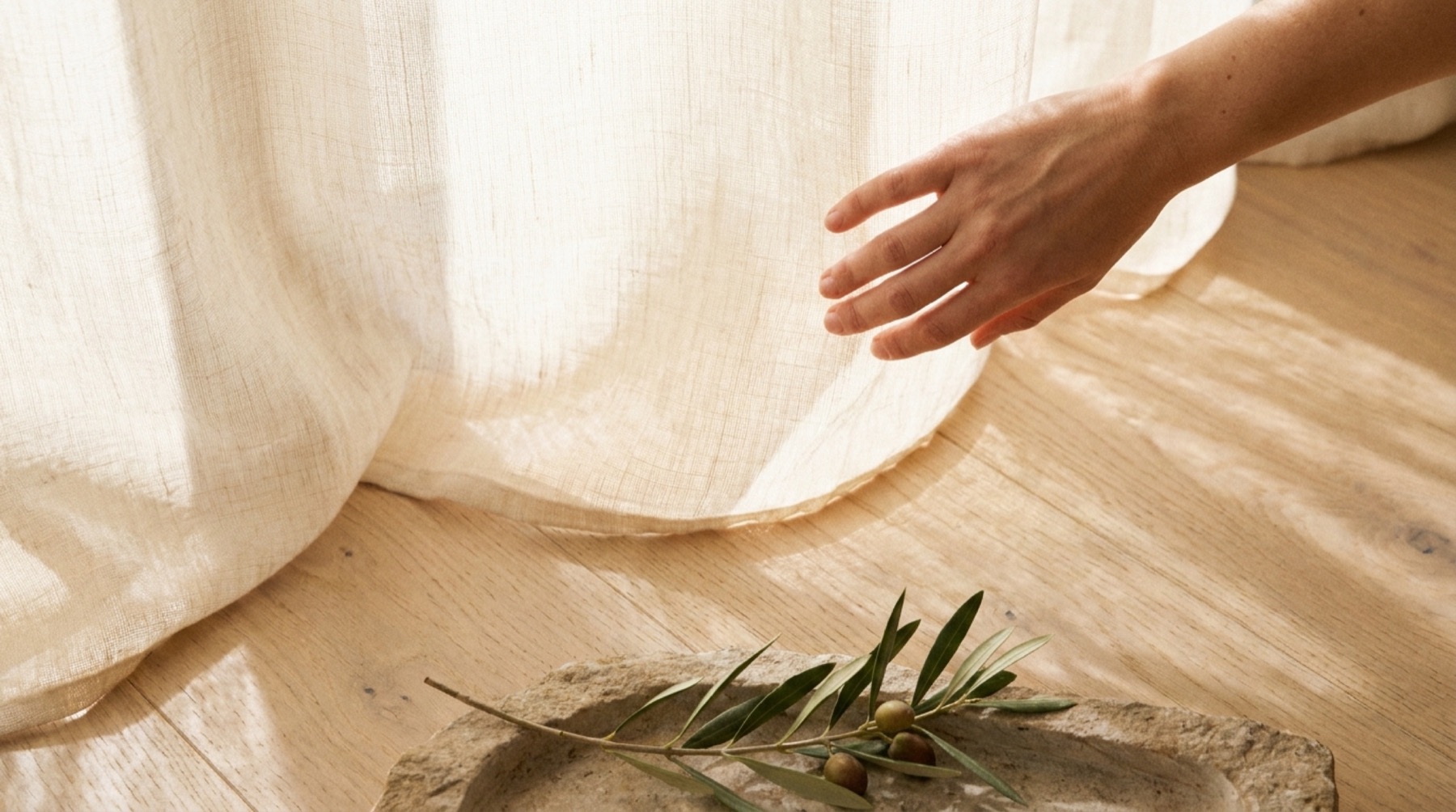 A hand reaching to a sheer linen curtain in low sun, an olive sprig resting in a stone dish on an oak floor